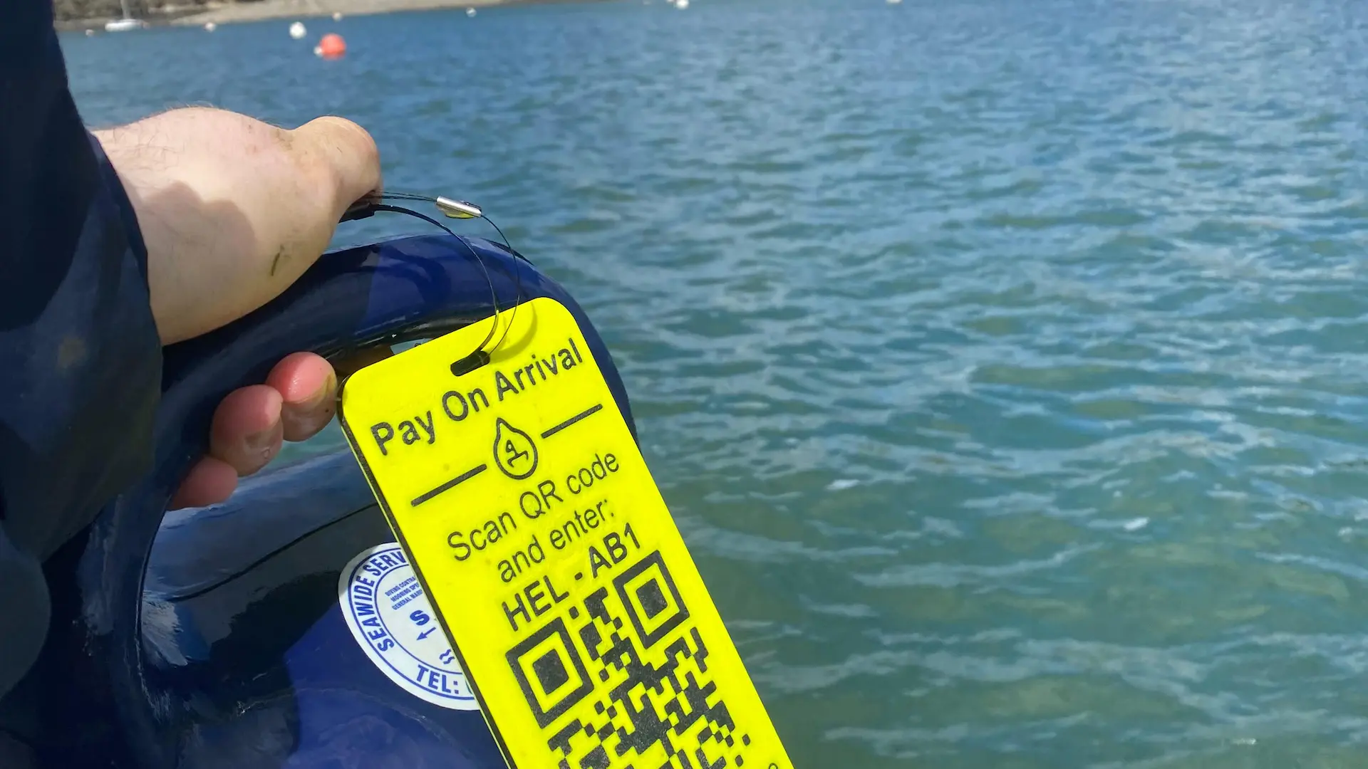 A yellow Pay On Arrival tag reading 'Scan QR code and enter: HEL-AB1' clipped to a mooring buoy pickup, with the harbour behind it